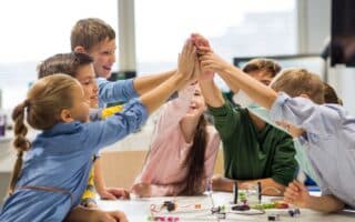 A group of six smiling children high-five each other over a table with electronics and wires, in a bright classroom. The kids appear happy and engaged, suggesting teamwork and collaboration on a science or STEM project.