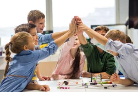 A group of six smiling children high-five each other over a table with electronics and wires, in a bright classroom. The kids appear happy and engaged, suggesting teamwork and collaboration on a science or STEM project.