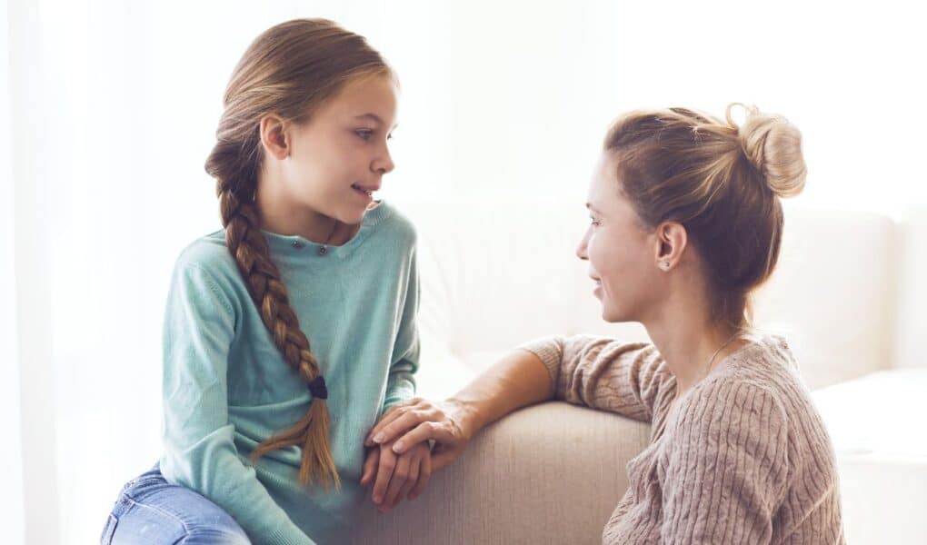 A woman and a young girl sit facing each other indoors. The woman, with her hair in a bun, smiles warmly at the girl, who has a long plait and wears a teal jumper. They hold hands and appear to be having a supportive conversation on a light sofa.