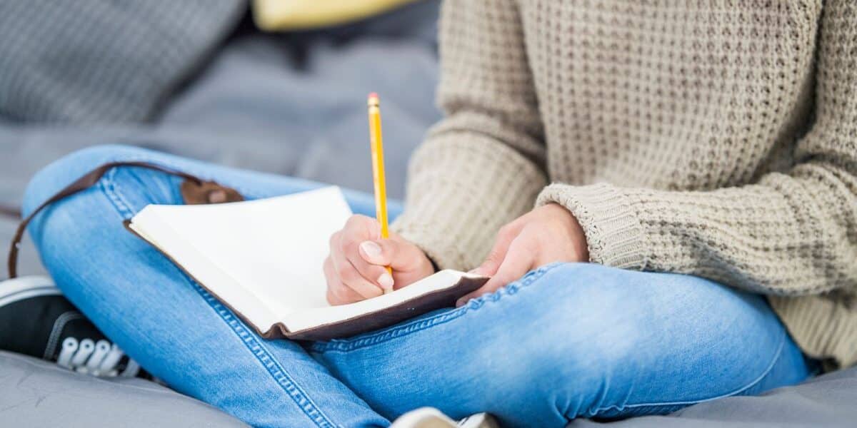 A person in a beige knitted jumper and blue jeans sits cross-legged on a bed, writing in a notebook with a yellow pencil. The person's face is not visible. Cushions and a grey blanket are in the background, creating a cosy setting.