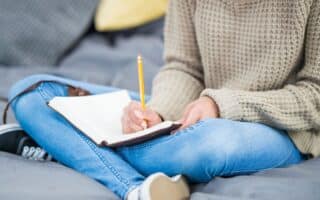 A person in a beige knitted jumper and blue jeans sits cross-legged on a bed, writing in a notebook with a yellow pencil. The person's face is not visible. Cushions and a grey blanket are in the background, creating a cosy setting.