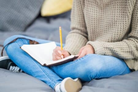 A person in a beige knitted jumper and blue jeans sits cross-legged on a bed, writing in a notebook with a yellow pencil. The person's face is not visible. Cushions and a grey blanket are in the background, creating a cosy setting.
