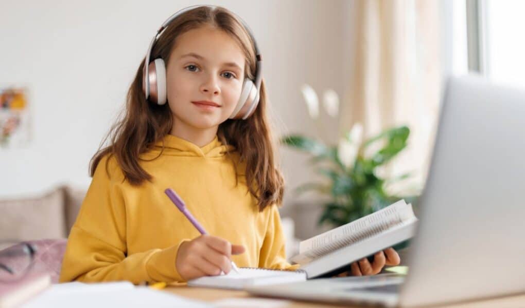 A young girl in a yellow hoodie wears large headphones and sits at a desk, writing in a notebook while looking at a laptop. She appears focused, with an open book in one hand. The background shows a bright room with plants and a window.