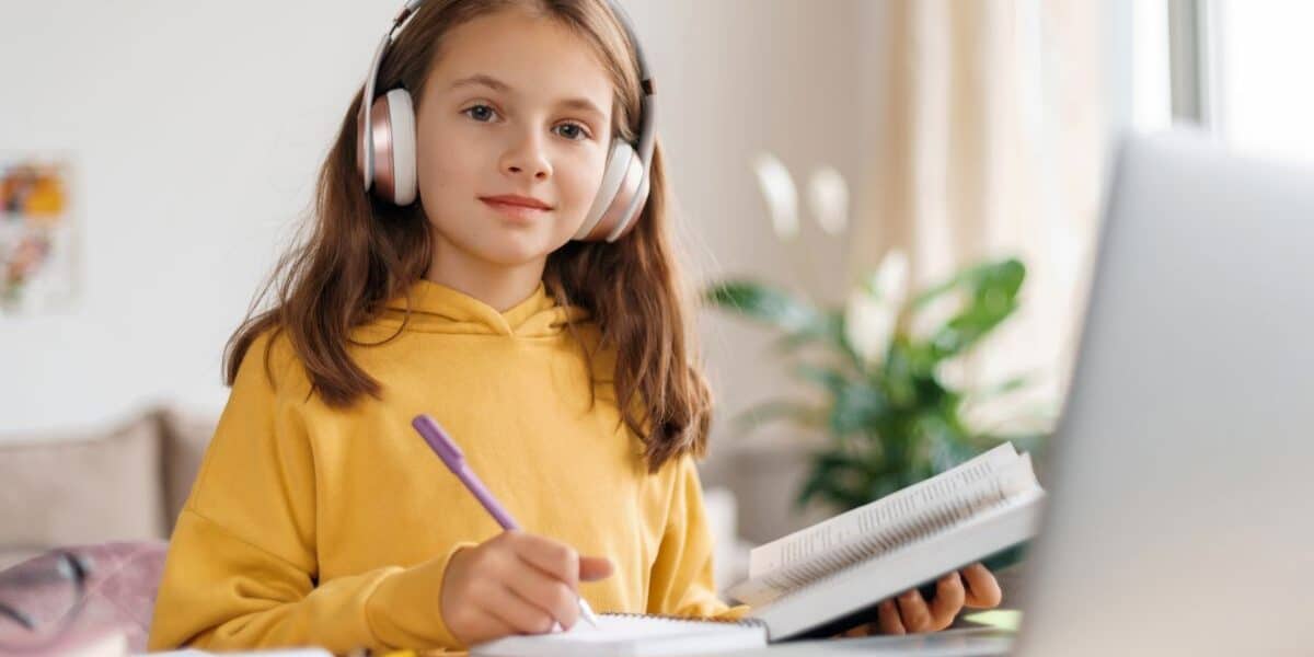 A young girl in a yellow hoodie wears large headphones and sits at a desk, writing in a notebook while looking at a laptop. She appears focused, with an open book in one hand. The background shows a bright room with plants and a window.