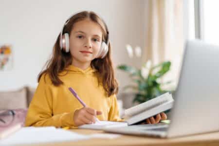 A young girl in a yellow hoodie wears large headphones and sits at a desk, writing in a notebook while looking at a laptop. She appears focused, with an open book in one hand. The background shows a bright room with plants and a window.
