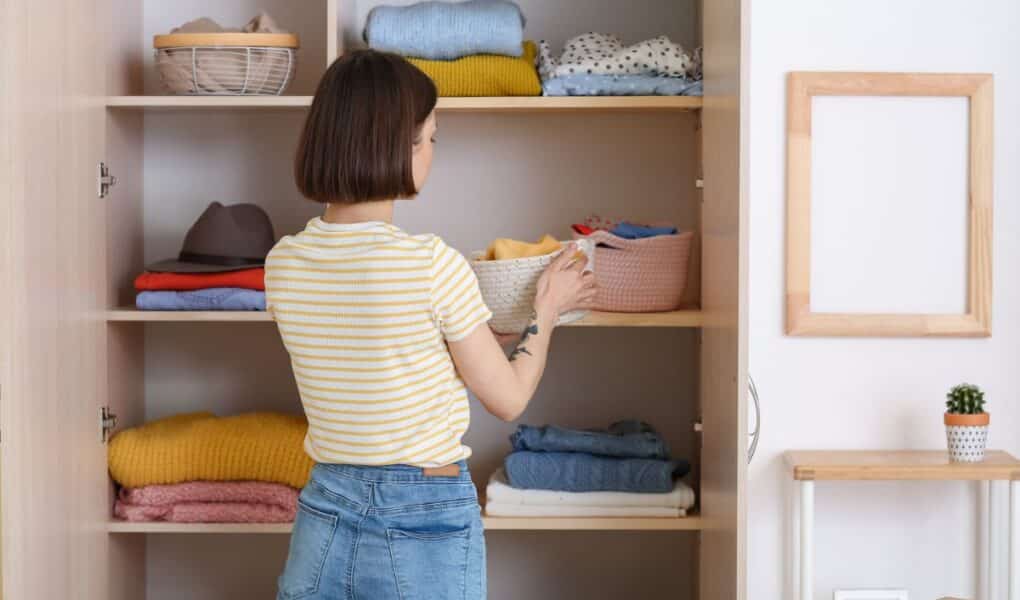 A woman with short brown hair, wearing a yellow striped t-shirt and blue jeans, organises folded clothes and baskets on shelves in an open wardrobe. Some shelves have neatly stacked jumpers and shirts. A small potted cactus sits nearby.