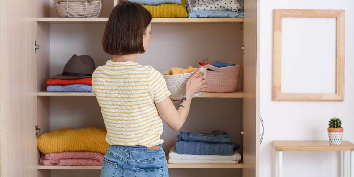 A woman with short brown hair, wearing a yellow striped t-shirt and blue jeans, organises folded clothes and baskets on shelves in an open wardrobe. Some shelves have neatly stacked jumpers and shirts. A small potted cactus sits nearby.