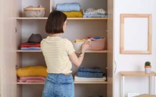 A woman with short brown hair, wearing a yellow striped t-shirt and blue jeans, organises folded clothes and baskets on shelves in an open wardrobe. Some shelves have neatly stacked jumpers and shirts. A small potted cactus sits nearby.