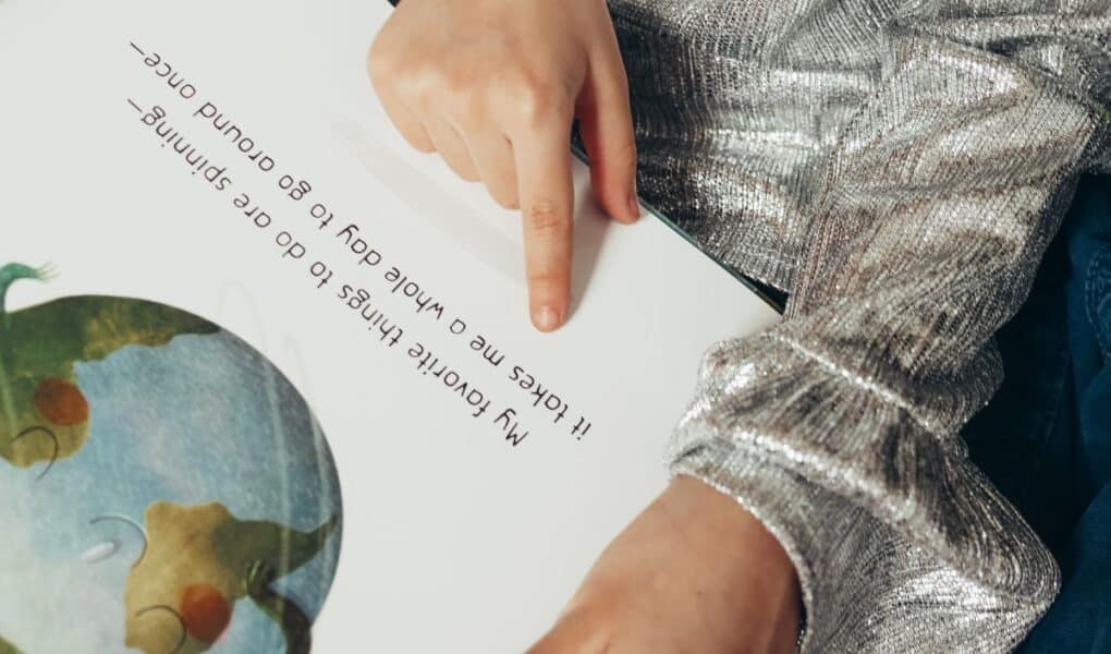 A child wearing a shiny silver long-sleeve top sits and points to text in a picture book. The text says, “My favourite things to do are spinning— It takes me a whole day to go round once—”. An illustration of Earth appears on the left page.