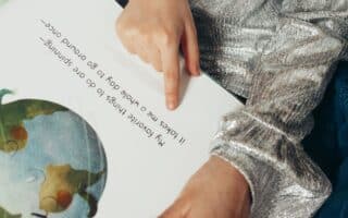A child wearing a shiny silver long-sleeve top sits and points to text in a picture book. The text says, “My favourite things to do are spinning— It takes me a whole day to go round once—”. An illustration of Earth appears on the left page.