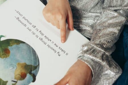 A child wearing a shiny silver long-sleeve top sits and points to text in a picture book. The text says, “My favourite things to do are spinning— It takes me a whole day to go round once—”. An illustration of Earth appears on the left page.