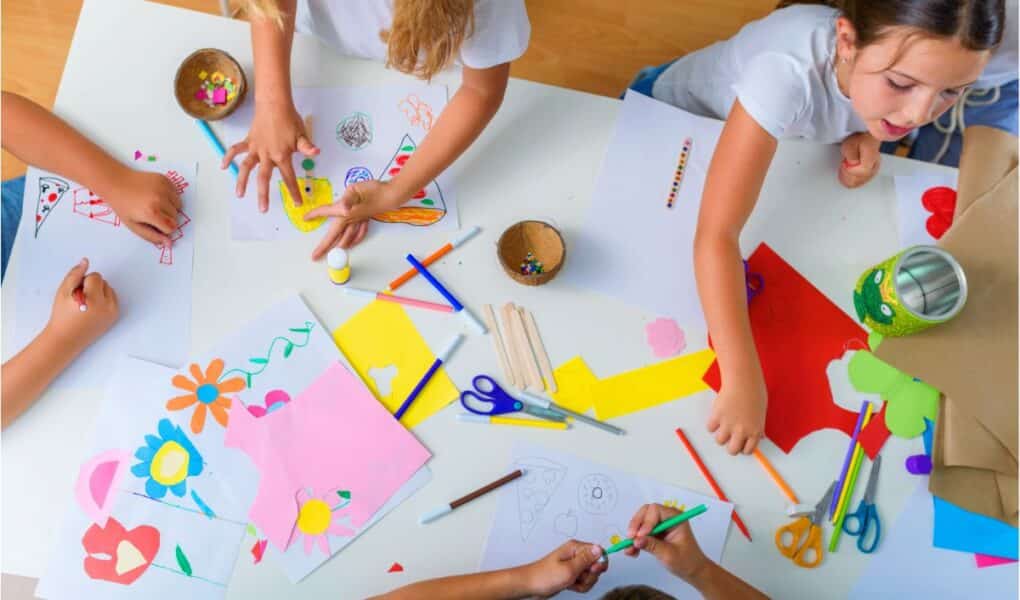 Four children sit and stand around a white table covered with colourful craft supplies, paper cut-outs, felt-tip pens, glue, and scissors. They draw and create art, surrounded by drawings of flowers and shapes on white and coloured paper. The setting is bright and playful.