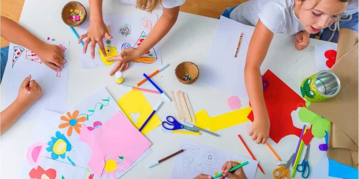 Four children sit and stand around a white table covered with colourful craft supplies, paper cut-outs, felt-tip pens, glue, and scissors. They draw and create art, surrounded by drawings of flowers and shapes on white and coloured paper. The setting is bright and playful.