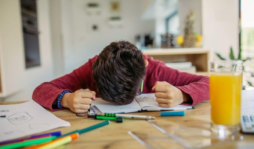 A child with brown hair wearing a red jumper sits at a table with his head down on an open notebook, fists clenched in frustration. Colourful felt-tip pens, papers, and a glass of orange juice are on the table in a bright, modern room.