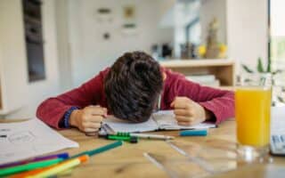 A child with brown hair wearing a red jumper sits at a table with his head down on an open notebook, fists clenched in frustration. Colourful felt-tip pens, papers, and a glass of orange juice are on the table in a bright, modern room.