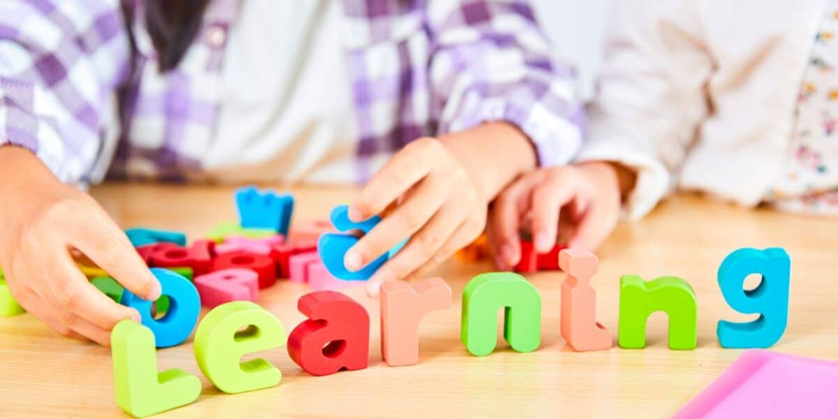 Two children arrange colourful foam letters spelling "Learning" on a wooden table. Their hands are visible, and they wear checked and white shirts. Other foam letters, coloured pencils, and paper can be seen in the background.