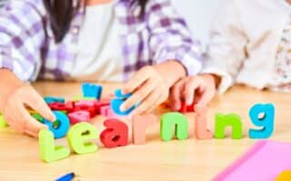 Two children arrange colourful foam letters spelling "Learning" on a wooden table. Their hands are visible, and they wear checked and white shirts. Other foam letters, coloured pencils, and paper can be seen in the background.