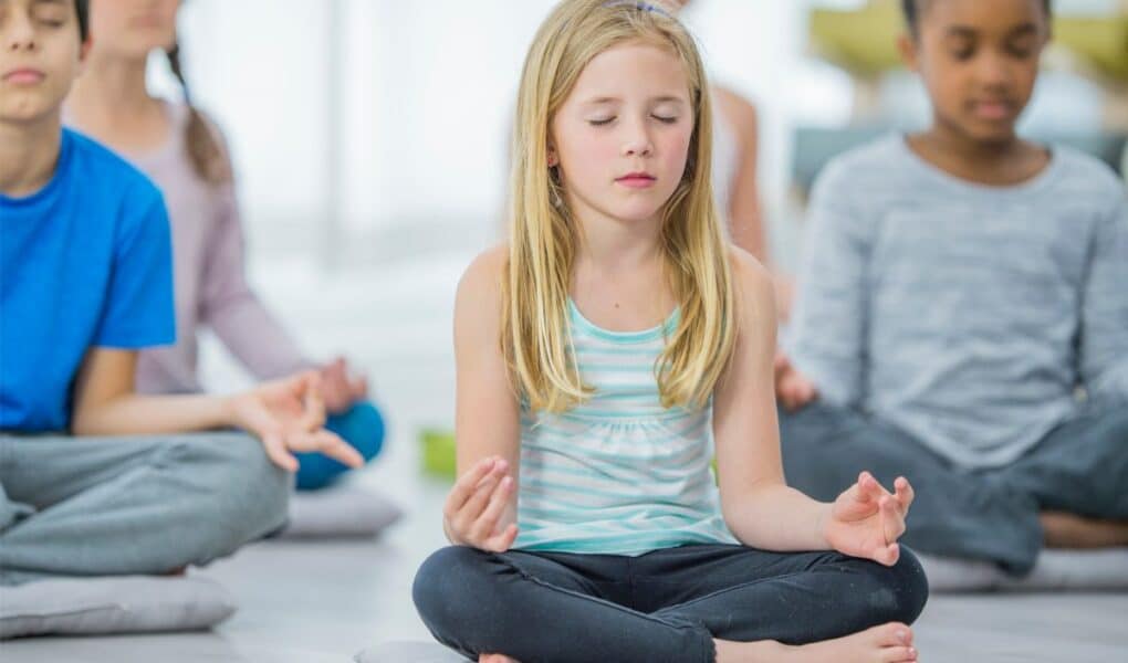 A group of children sit cross-legged on cushions indoors, meditating with eyes closed and hands resting on their knees, fingers in a mudra. The focus is on a blonde girl in a striped vest in the centre, with other kids blurred in the background.