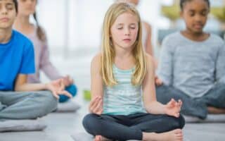 A group of children sit cross-legged on cushions indoors, meditating with eyes closed and hands resting on their knees, fingers in a mudra. The focus is on a blonde girl in a striped vest in the centre, with other kids blurred in the background.