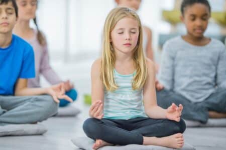 A group of children sit cross-legged on cushions indoors, meditating with eyes closed and hands resting on their knees, fingers in a mudra. The focus is on a blonde girl in a striped vest in the centre, with other kids blurred in the background.