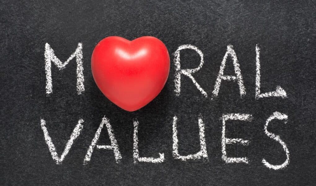 The words “MORAL VALUES” are written in white chalk on a blackboard. The letter “O” in “MORAL” is replaced by a red heart-shaped object, symbolising the importance of love and compassion in moral values.
