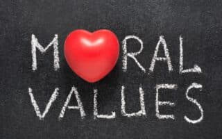 The words “MORAL VALUES” are written in white chalk on a blackboard. The letter “O” in “MORAL” is replaced by a red heart-shaped object, symbolising the importance of love and compassion in moral values.