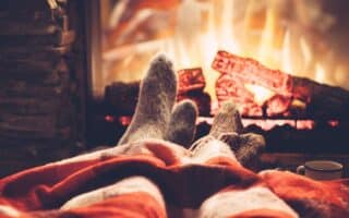 Two people with woollen socks relax by a roaring fireplace, their legs covered by a red-and-white checked blanket. Logs burn brightly in the hearth, and a white mug sits on the floor nearby, evoking warmth and cosiness indoors.