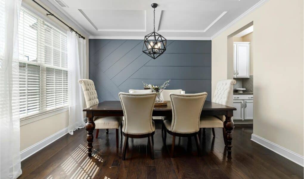 Modern dining room with dark wood floor, dark wood table, and six beige upholstered chairs. The back wall is painted navy with a geometric pattern. White ceiling with a black geometric pendant light. Natural light streams in through large windows with white curtains.