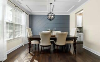 Modern dining room with dark wood floor, dark wood table, and six beige upholstered chairs. The back wall is painted navy with a geometric pattern. White ceiling with a black geometric pendant light. Natural light streams in through large windows with white curtains.