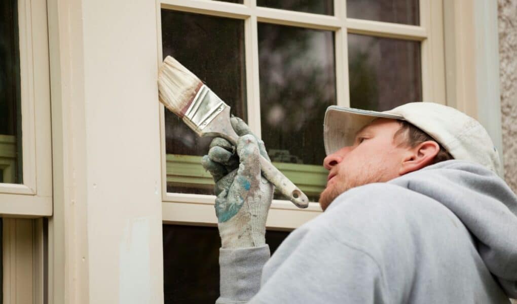 A person wearing a grey hoodie, white cap, and paint-stained glove carefully paints the wooden trim of a window with a paintbrush. The person is focused on painting along the edge, with glass window panes and a textured wall visible.