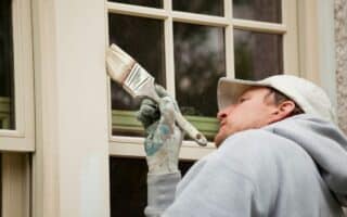 A person wearing a grey hoodie, white cap, and paint-stained glove carefully paints the wooden trim of a window with a paintbrush. The person is focused on painting along the edge, with glass window panes and a textured wall visible.