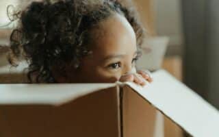 A young child with curly hair peers over the edge of a large cardboard box. Only the top half of the child's face and hands are visible, giving a sense of curiosity and playfulness. The scene is softly lit, creating a warm, gentle atmosphere.