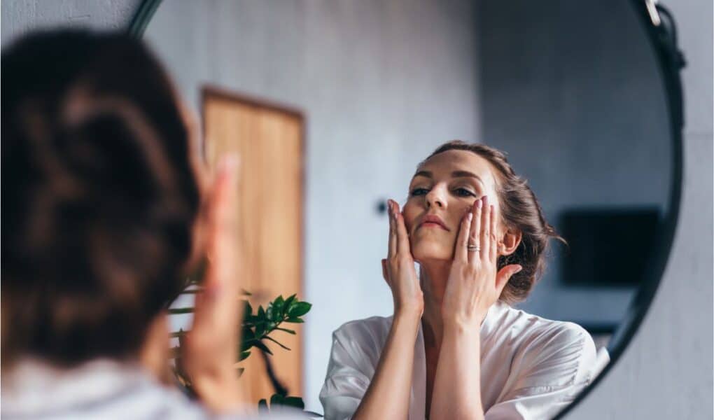 A woman in a white dressing gown stands in front of a round mirror, gently touching her face with both hands. Her hair is tied back, and the room has soft lighting with a plant visible in the background. She appears to be following a skincare routine.