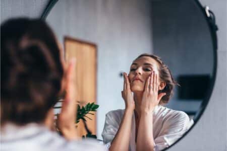 A woman in a white dressing gown stands in front of a round mirror, gently touching her face with both hands. Her hair is tied back, and the room has soft lighting with a plant visible in the background. She appears to be following a skincare routine.