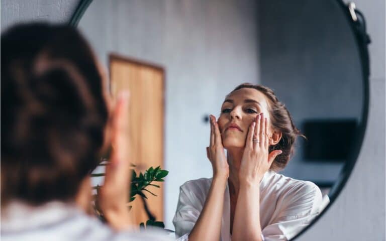 A woman in a white dressing gown stands in front of a round mirror, gently touching her face with both hands. Her hair is tied back, and the room has soft lighting with a plant visible in the background. She appears to be following a skincare routine.
