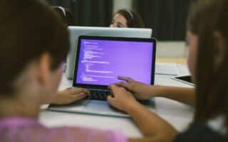 Two girls sit at a table, working together on a laptop displaying lines of code on a purple screen. One girl types while the other points to the screen. Additional laptops and two other children are partially visible in the background.