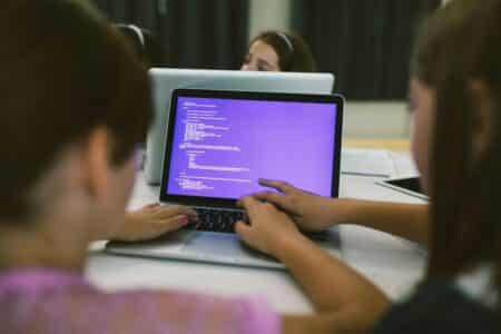 Two girls sit at a table, working together on a laptop displaying lines of code on a purple screen. One girl types while the other points to the screen. Additional laptops and two other children are partially visible in the background.