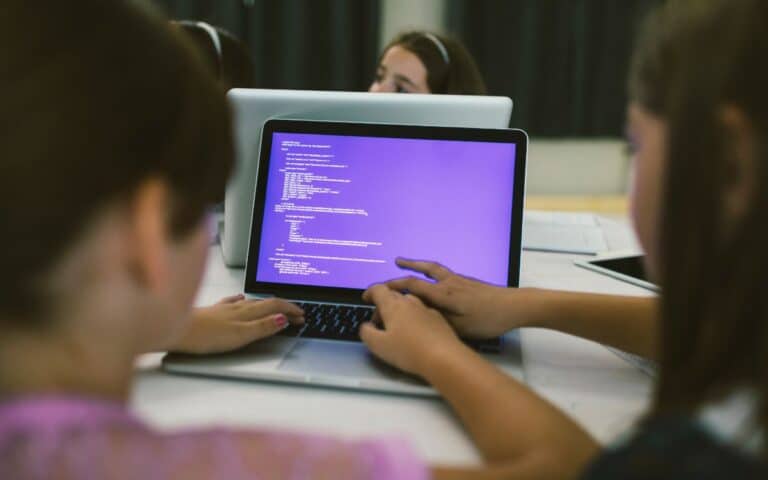 Two girls sit at a table, working together on a laptop displaying lines of code on a purple screen. One girl types while the other points to the screen. Additional laptops and two other children are partially visible in the background.