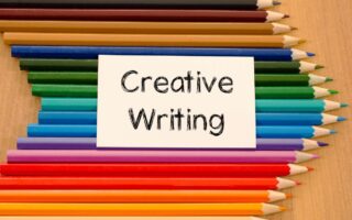 A white card with "Creative Writing" in black text is centred on a wooden surface, surrounded by neatly arranged coloured pencils in various shades, pointing inward from the top and bottom edges towards the card.