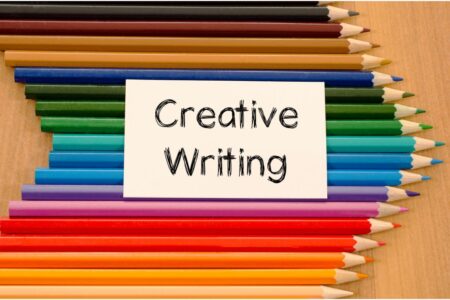 A white card with "Creative Writing" in black text is centred on a wooden surface, surrounded by neatly arranged coloured pencils in various shades, pointing inward from the top and bottom edges towards the card.