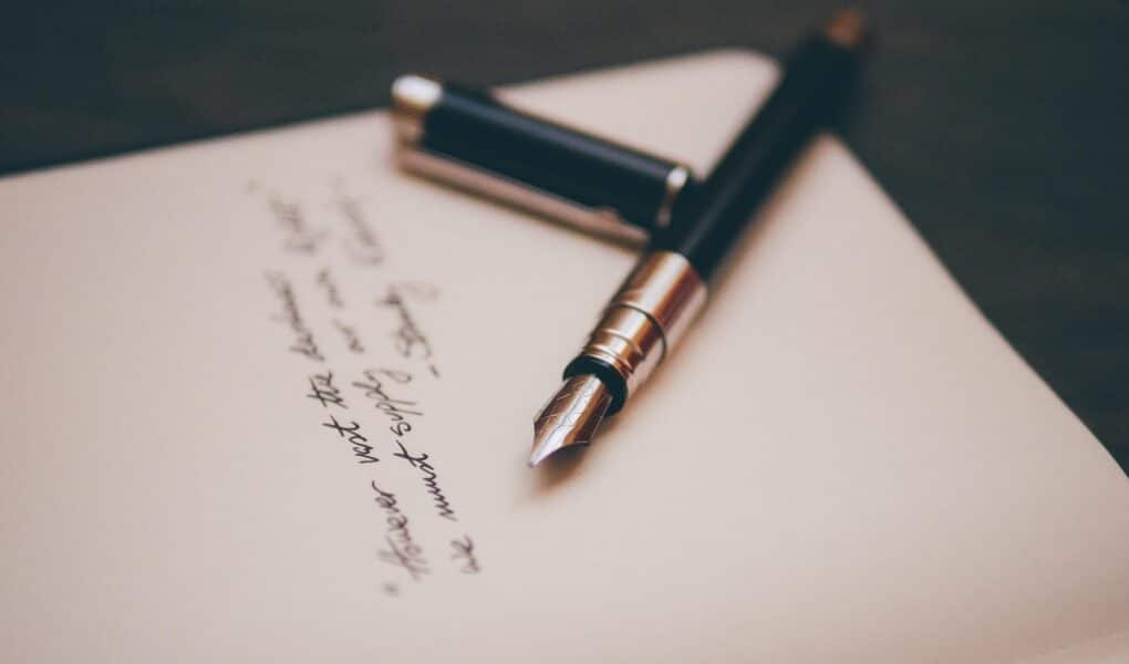 A black fountain pen with a silver clip rests on an open notebook. The cap is off and lies next to the pen. There is handwritten text in black ink on the page, slightly out of focus, with the pen tip pointing towards the writing.