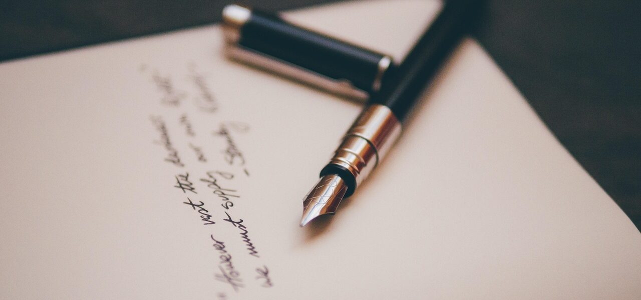 A black fountain pen with a silver clip rests on an open notebook. The cap is off and lies next to the pen. There is handwritten text in black ink on the page, slightly out of focus, with the pen tip pointing towards the writing.