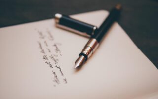 A black fountain pen with a silver clip rests on an open notebook. The cap is off and lies next to the pen. There is handwritten text in black ink on the page, slightly out of focus, with the pen tip pointing towards the writing.