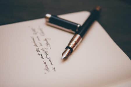 A black fountain pen with a silver clip rests on an open notebook. The cap is off and lies next to the pen. There is handwritten text in black ink on the page, slightly out of focus, with the pen tip pointing towards the writing.