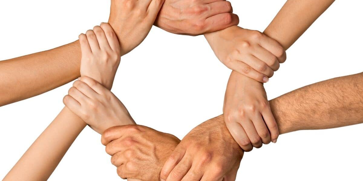 Eight hands form a circle by gripping each other’s wrists against a white background, symbolising unity, teamwork, and support. The hands vary in skin tones and sizes, suggesting diversity and co-operation among different people.