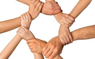 Eight hands form a circle by gripping each other’s wrists against a white background, symbolising unity, teamwork, and support. The hands vary in skin tones and sizes, suggesting diversity and co-operation among different people.