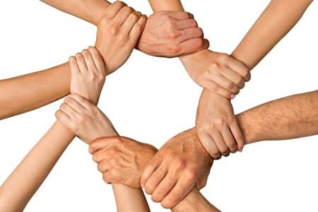 Eight hands form a circle by gripping each other’s wrists against a white background, symbolising unity, teamwork, and support. The hands vary in skin tones and sizes, suggesting diversity and co-operation among different people.