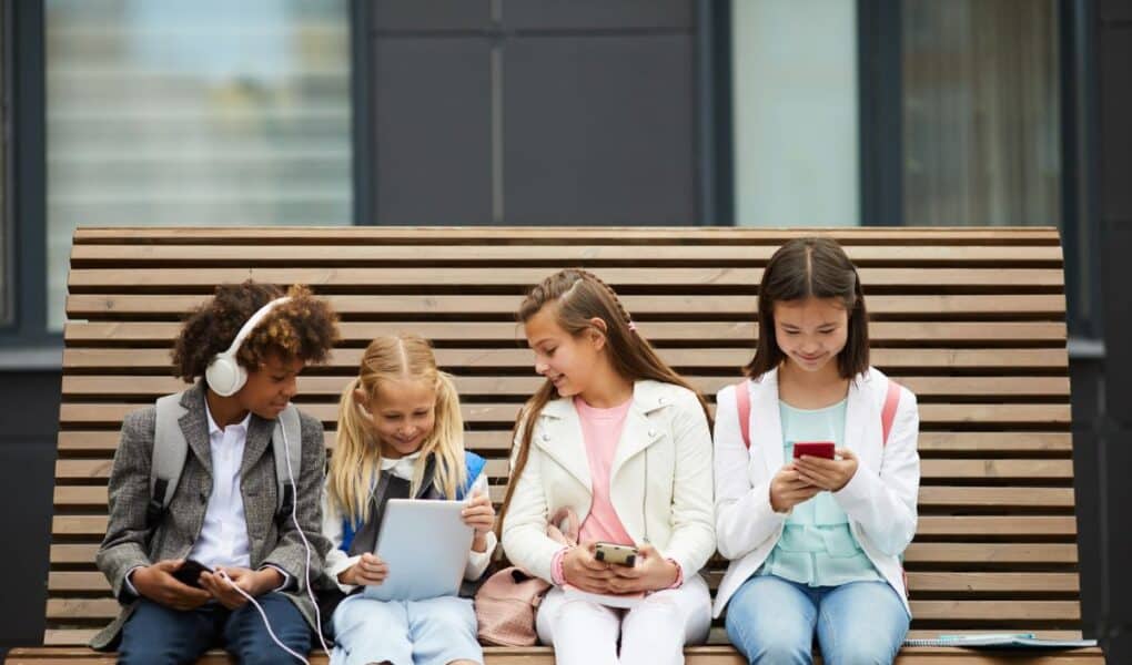 Four children sit on a wooden bench, each engaged with electronic devices. From left to right: a boy with headphones and a mobile, a girl with two plaits using a tablet, a girl with a mobile, and a girl in a white jacket looking at a smartphone.
