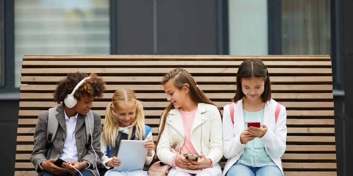 Four children sit on a wooden bench, each engaged with electronic devices. From left to right: a boy with headphones and a mobile, a girl with two plaits using a tablet, a girl with a mobile, and a girl in a white jacket looking at a smartphone.
