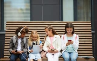 Four children sit on a wooden bench, each engaged with electronic devices. From left to right: a boy with headphones and a mobile, a girl with two plaits using a tablet, a girl with a mobile, and a girl in a white jacket looking at a smartphone.